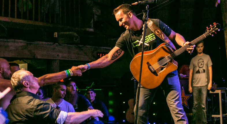 Man on stage reaching out to crowd, with a guitar